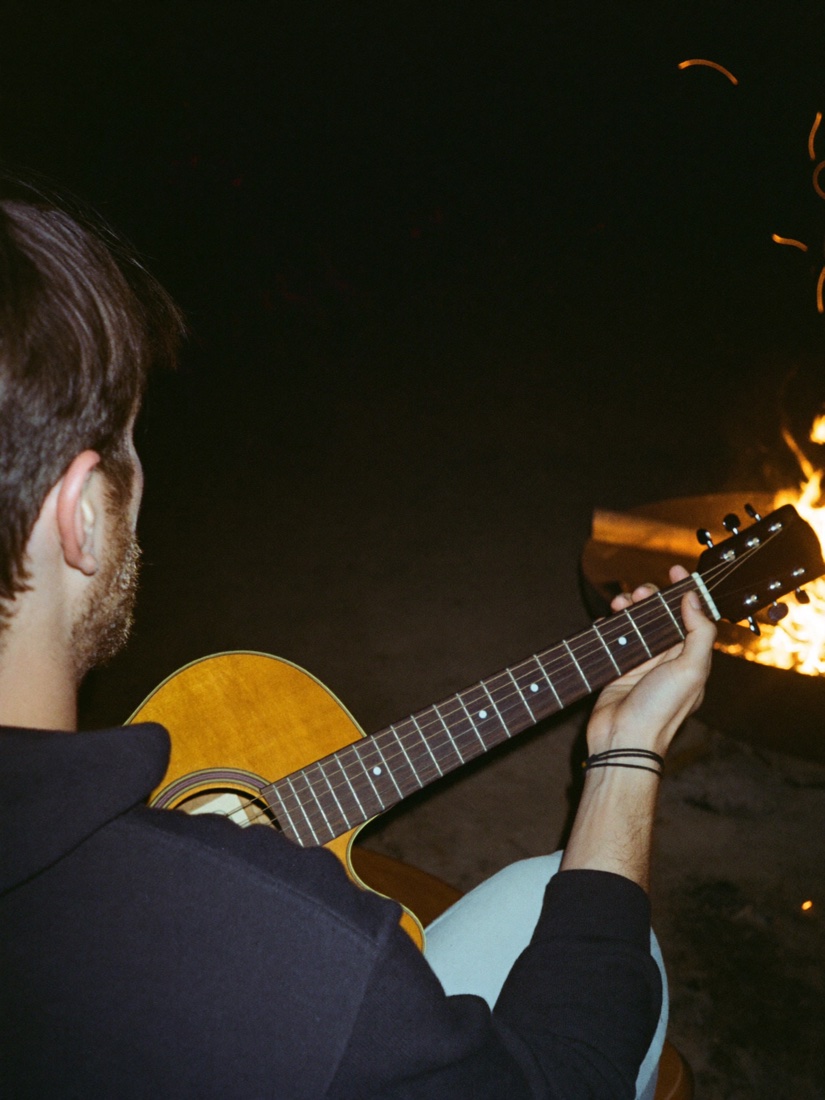 Over the shoulder of a man playing acoustic guitar beside the fire, sparks drifting in the dark