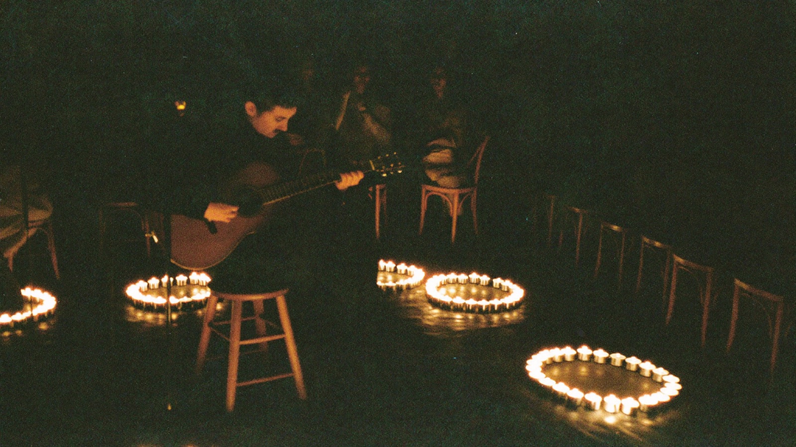 A guitarist on a stool ringed by tealights, playing to rows of bentwood chairs in a dark basement room
